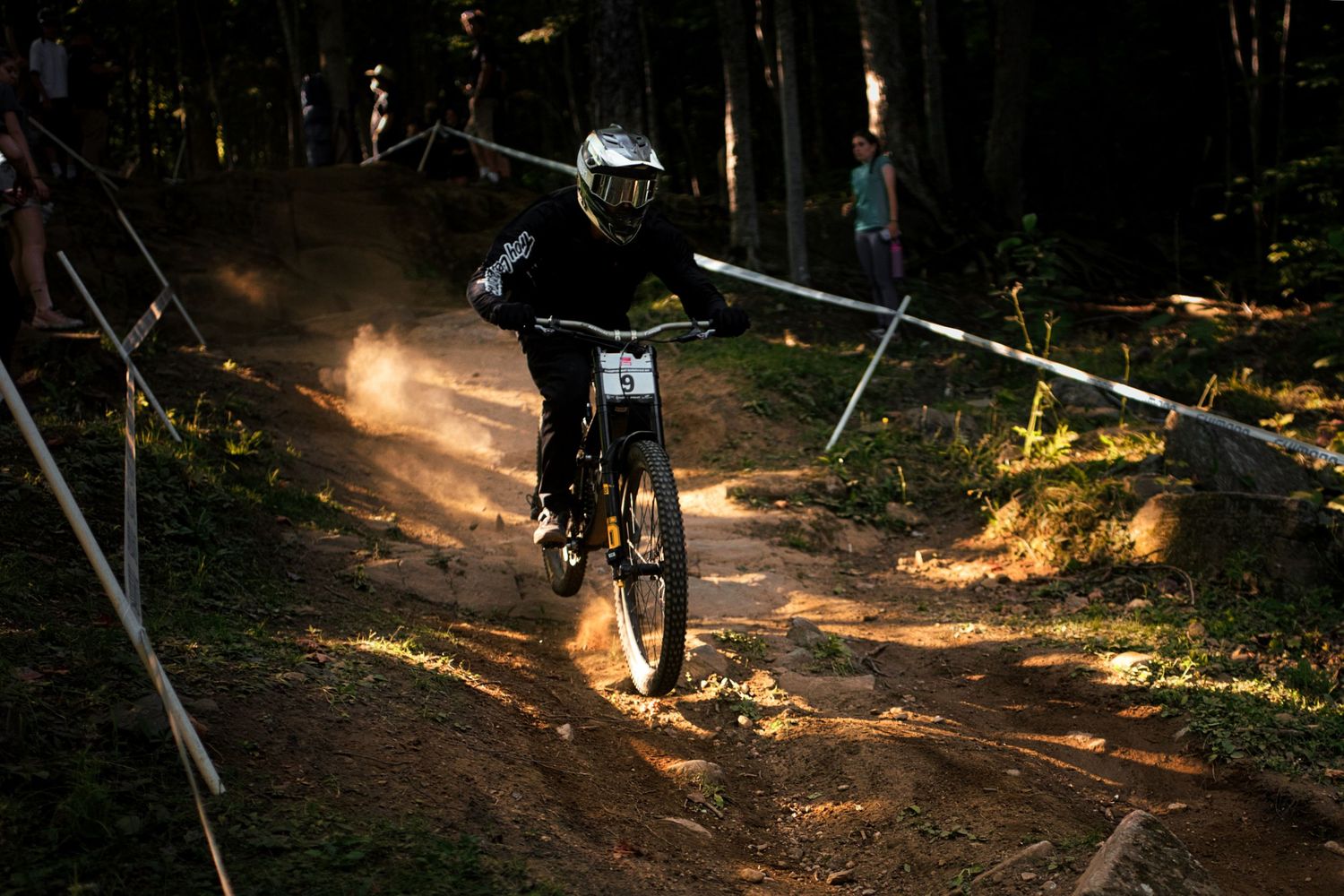 A mountain biker kicks up dust while descending a sunlit forest trail during a downhill competition.