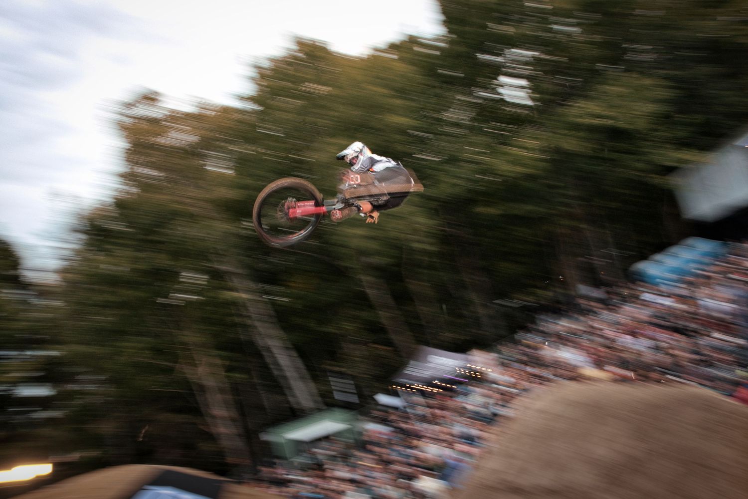 Mountain biker executing high-flying trick off dirt jump with motion-blurred crowd watching below by Dave Clément, Québec.