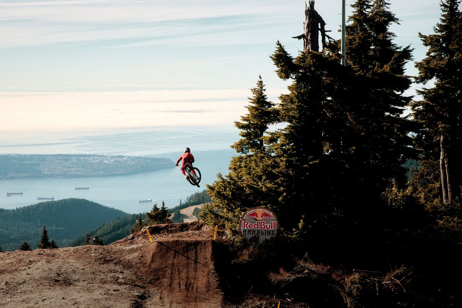Mountain biker soars off a dirt jump among evergreen trees with a sweeping coastal view and distant islands in the background.