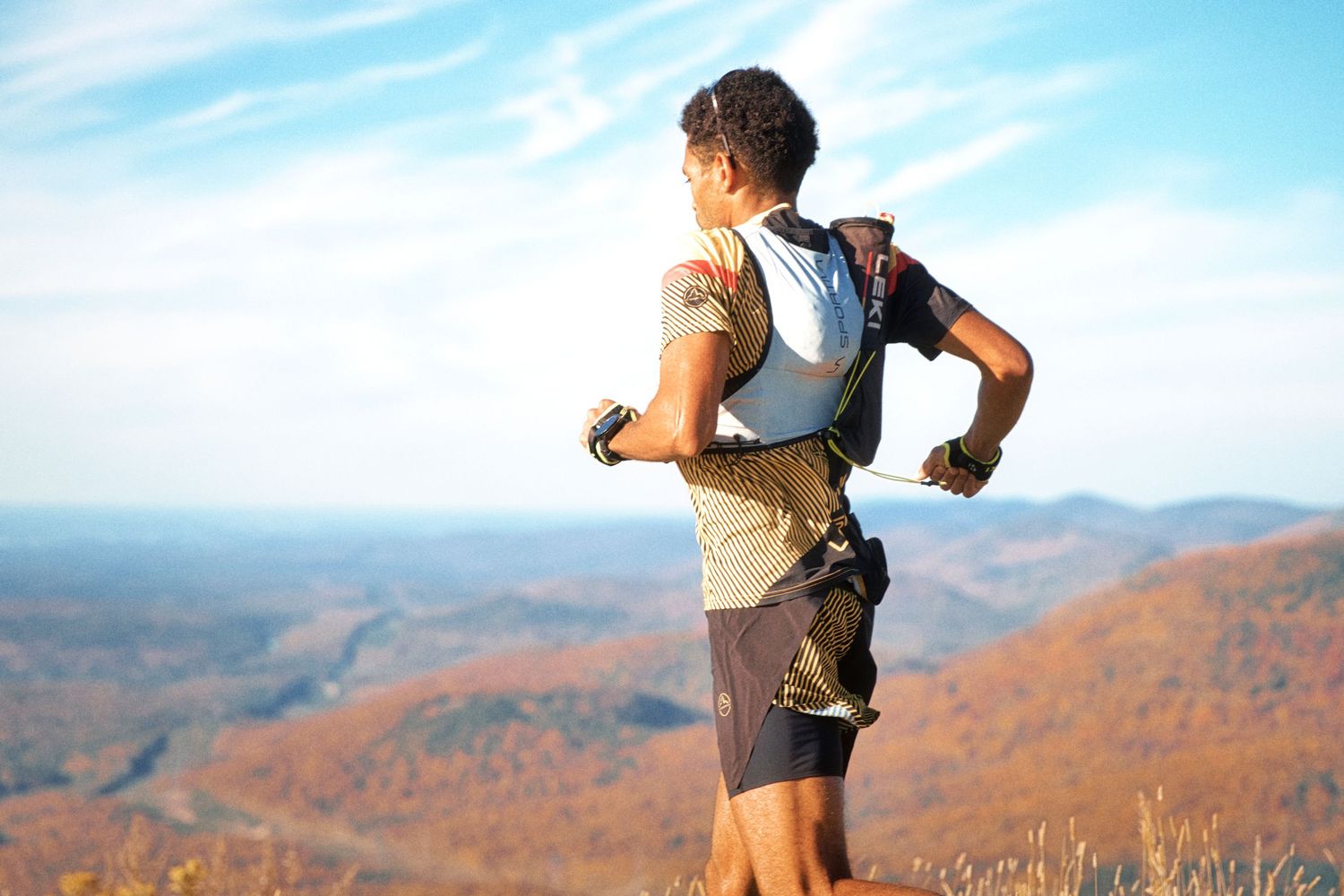 Trail runner with backpack races along mountain ridge with autumn foliage valley views under blue sky.