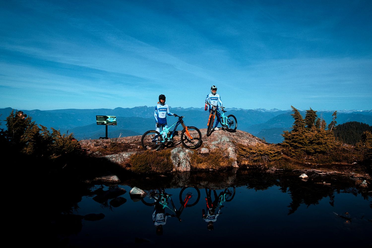 Two mountain bikers in helmets pause on a rocky alpine ledge above a still lake with reflections, overlooking distant blue mountain ranges.