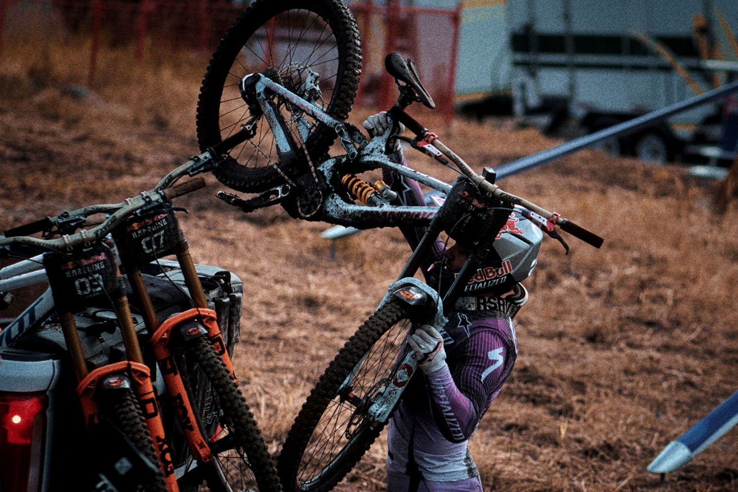 Mountain biker in a helmet and gloves lifts a full-suspension bike onto a truck-mounted bike rack on a dry grassy field at dusk.