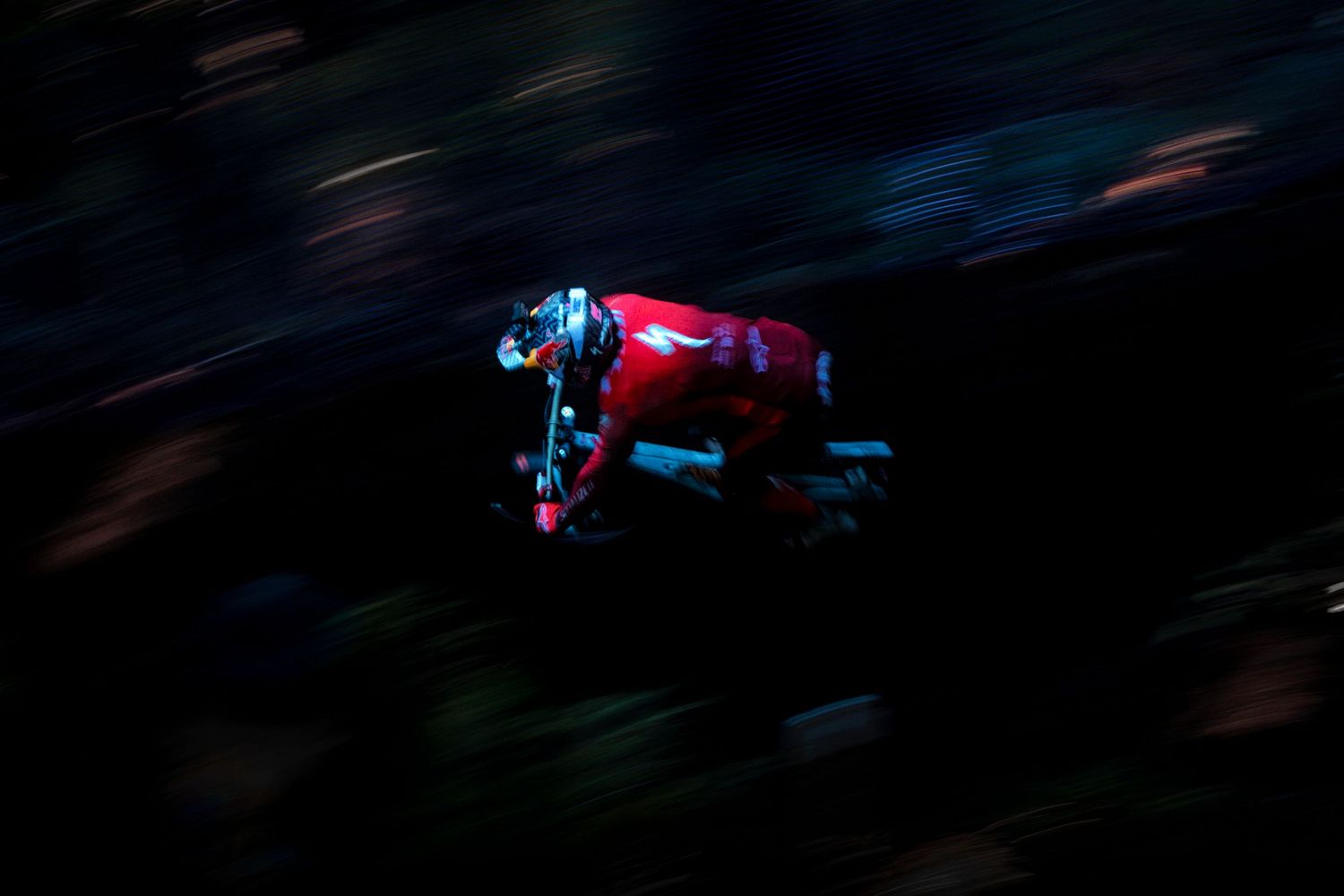 Mountain biker in a red jersey and helmet speeds downhill through a dark forest, captured in a dramatic panning shot with motion blur.