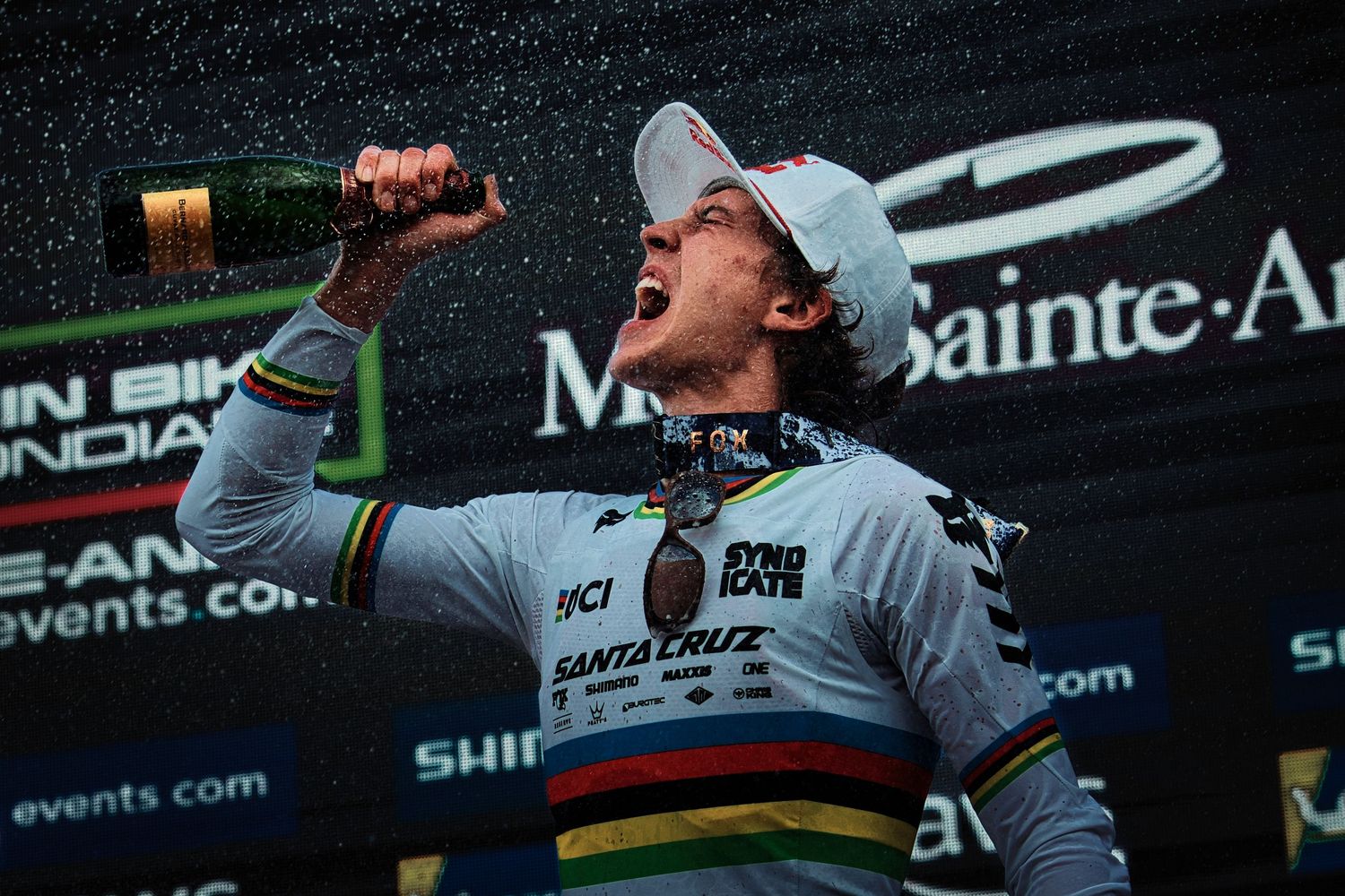 Cyclist celebrates victory on podium spraying champagne wearing world champion rainbow jersey and Santa Cruz cycling kit.