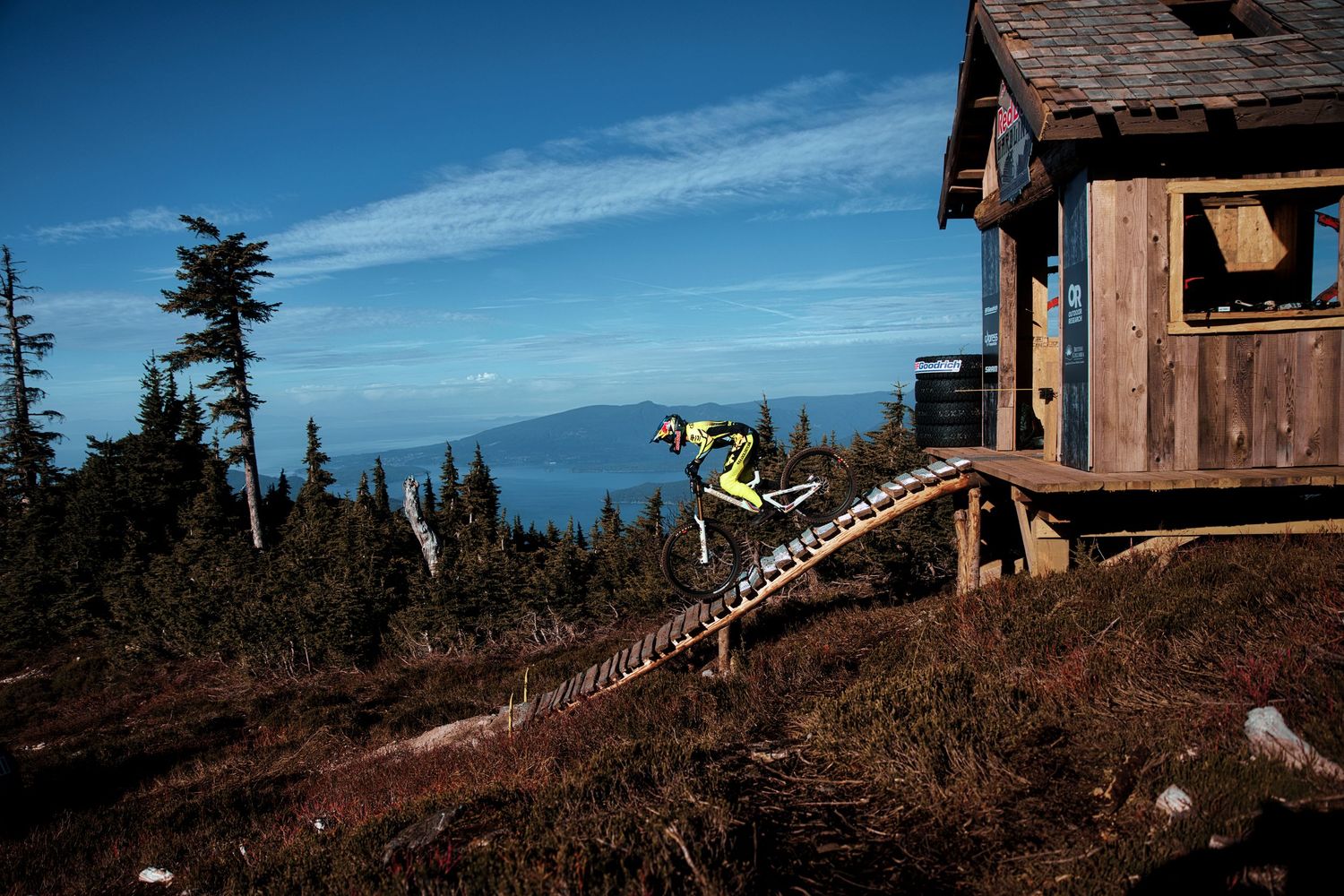 Mountain biker in yellow gear rides a wooden ramp beside a rustic cabin, with evergreen trees and distant ocean views in the background.