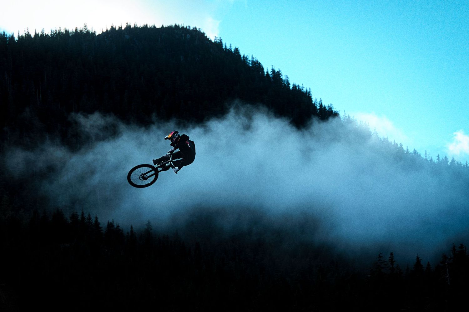 Mountain biker soars through the air above misty clouds with a dark forested hillside and blue sky in the background.