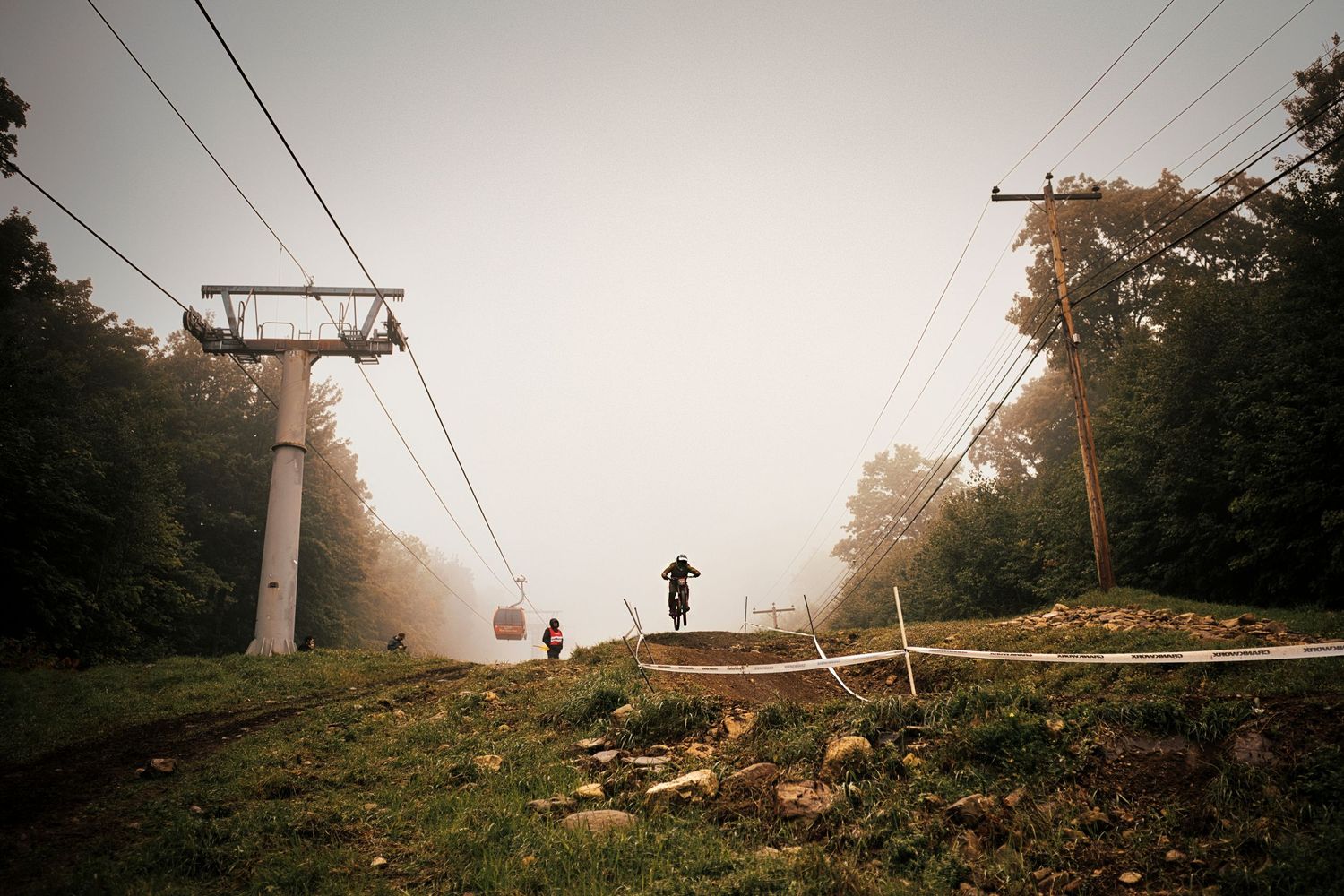 Mountain biker riding uphill on foggy ski slope with chairlift towers visible through misty trees.