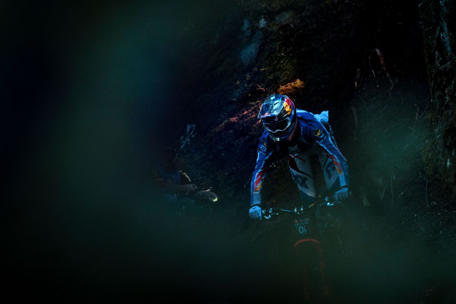 Mountain biker in a blue full-face helmet and riding gear speeds down a dark, shadowy forest trail.