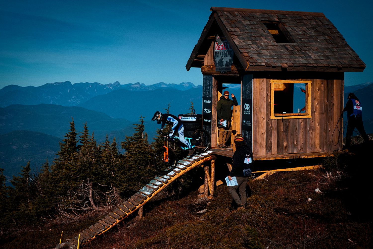 Mountain biker in full gear rides down a wooden ramp from a rustic start hut on a mountaintop with forested peaks in the background.
