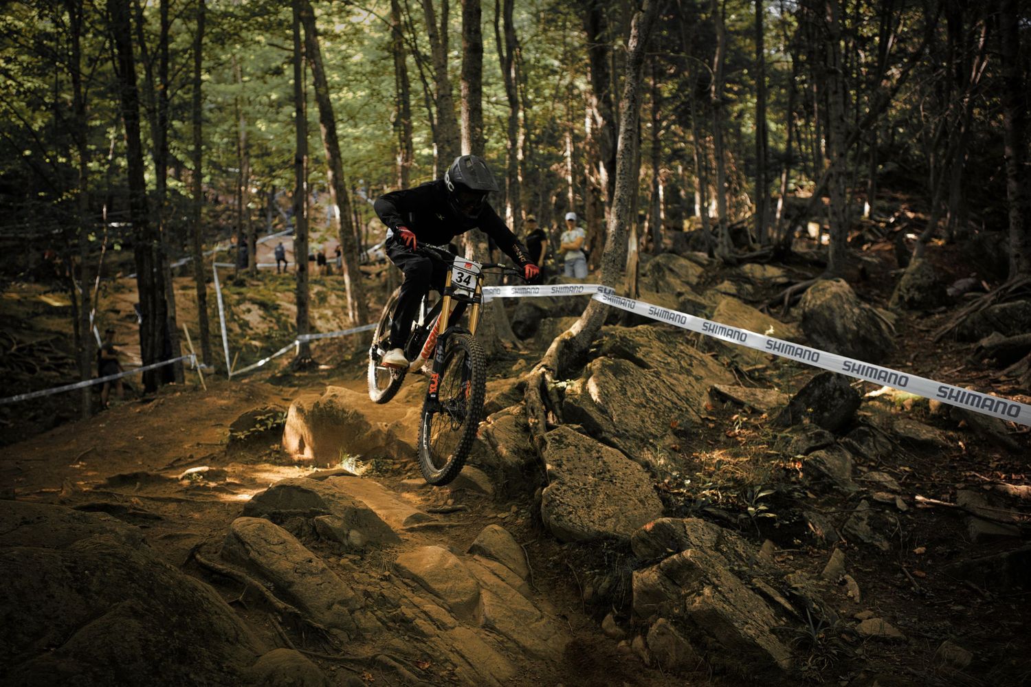 A mountain biker navigates a rocky technical section through a dense forest on a downhill race course by Dave Clément, Québec.
