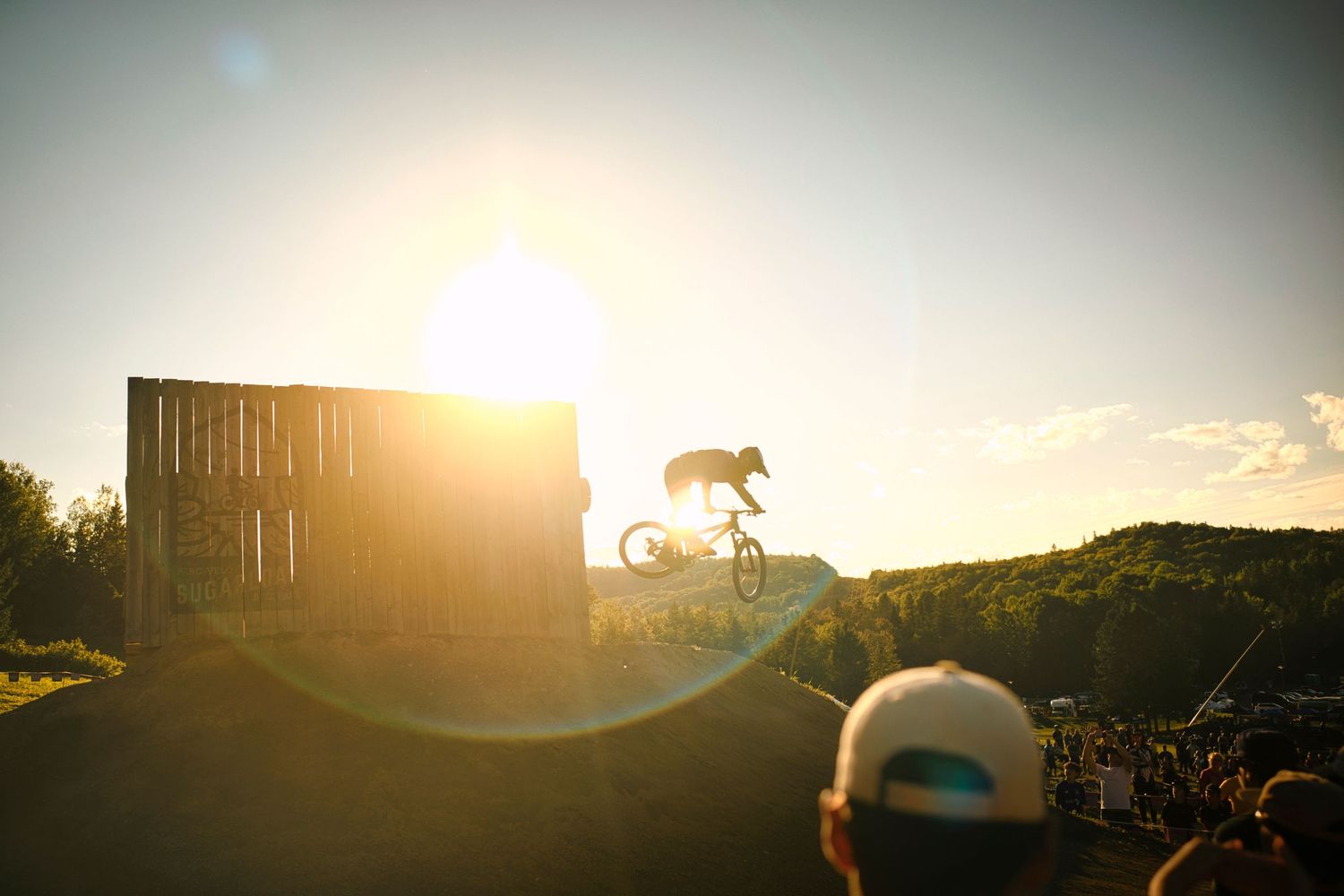 Silhouetted BMX rider performs a jump trick against a bright sunset sky at an outdoor bike park with spectators watching.