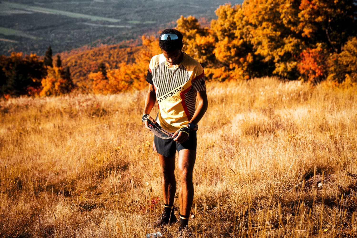 Trail runner in yellow vest navigating golden autumn grassland with colorful fall foliage hillside backdrop.