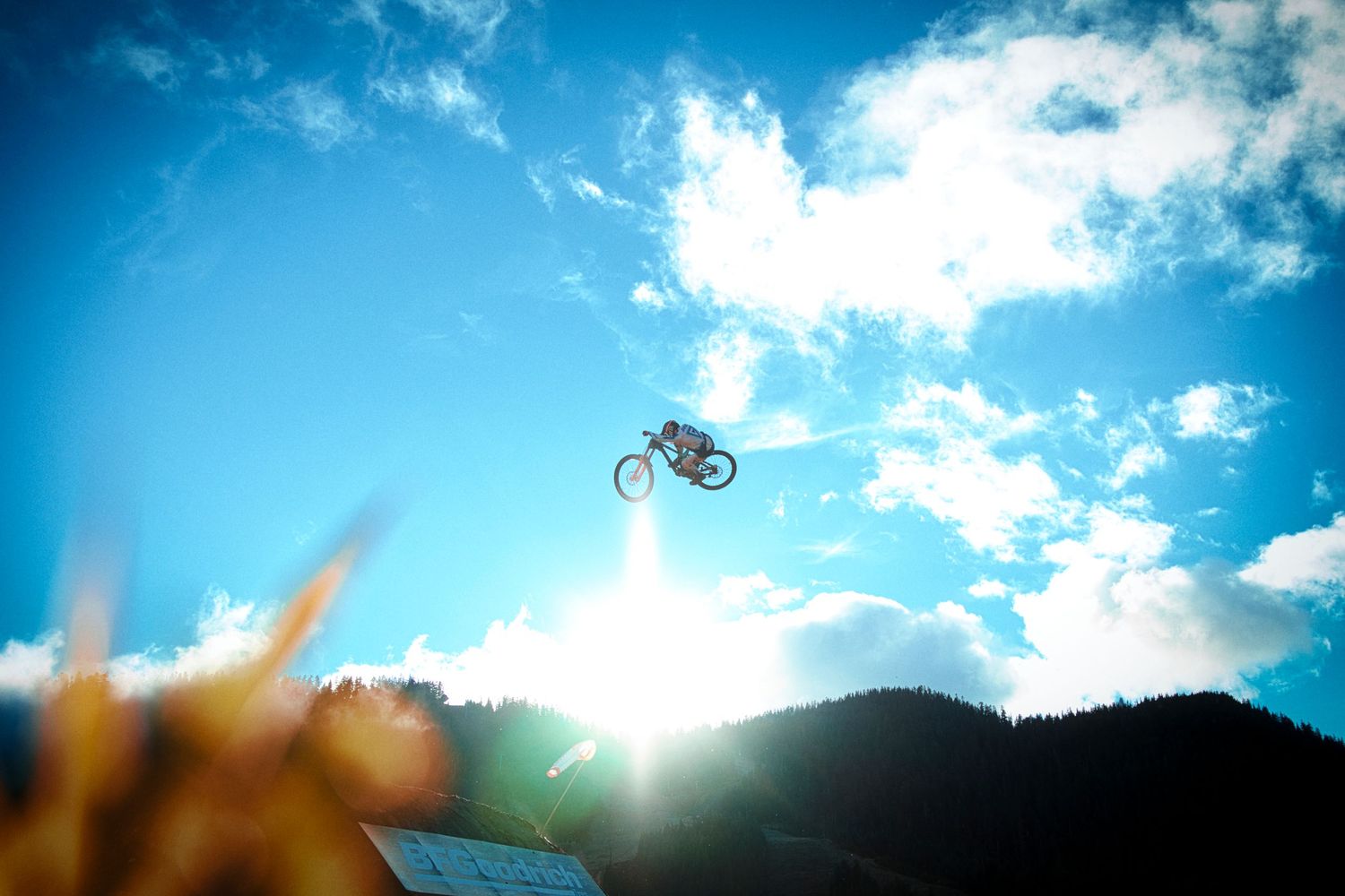 Mountain biker soars through a bright blue sky with clouds above a forested ridge, silhouetted against the glowing sun.