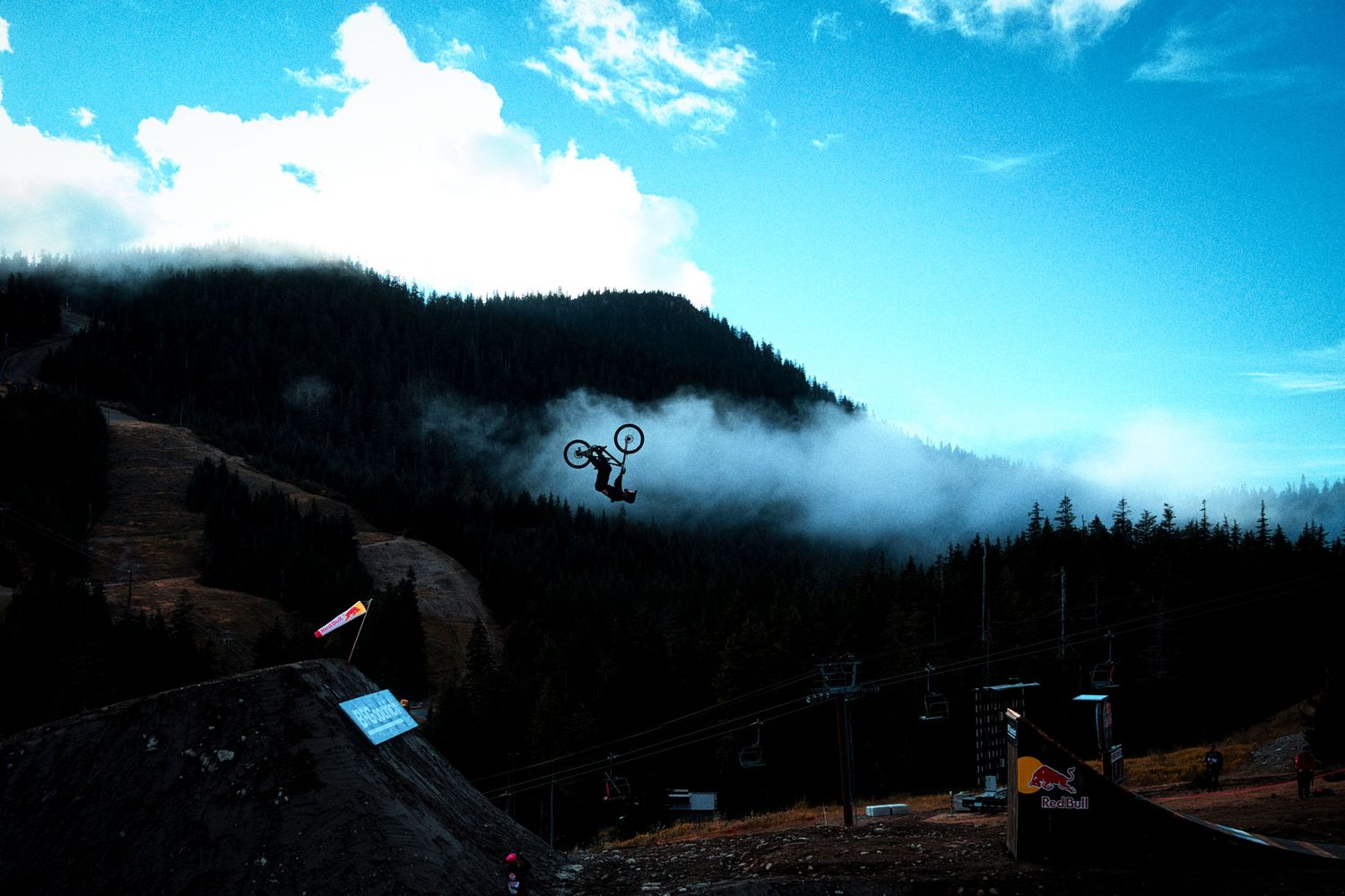 Mountain biker performs a airborne trick above a dirt jump with misty forested hills and a blue sky with clouds in the background.