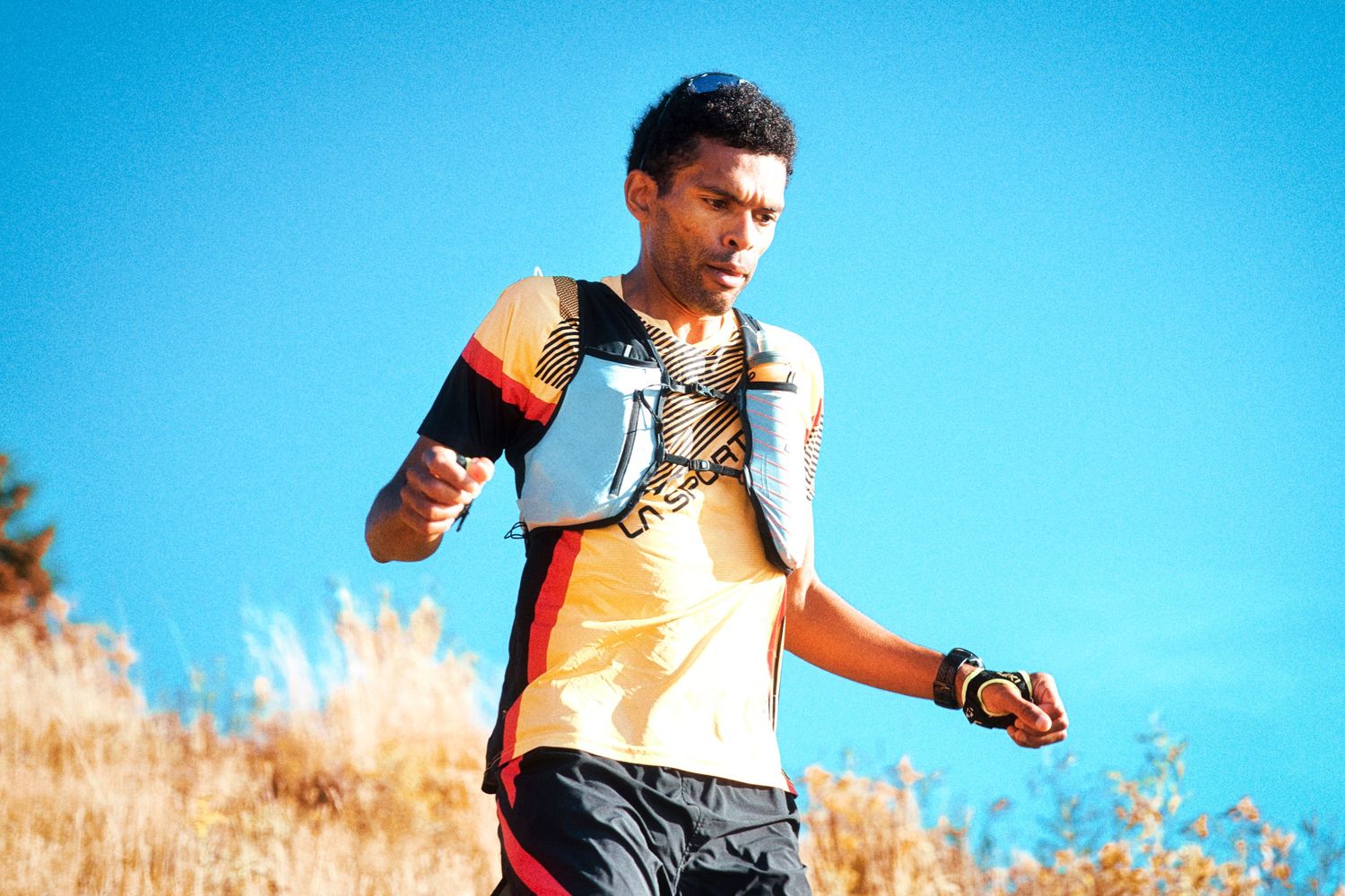Male trail runner in colorful vest navigating a sunny hillside during an outdoor mountain race.