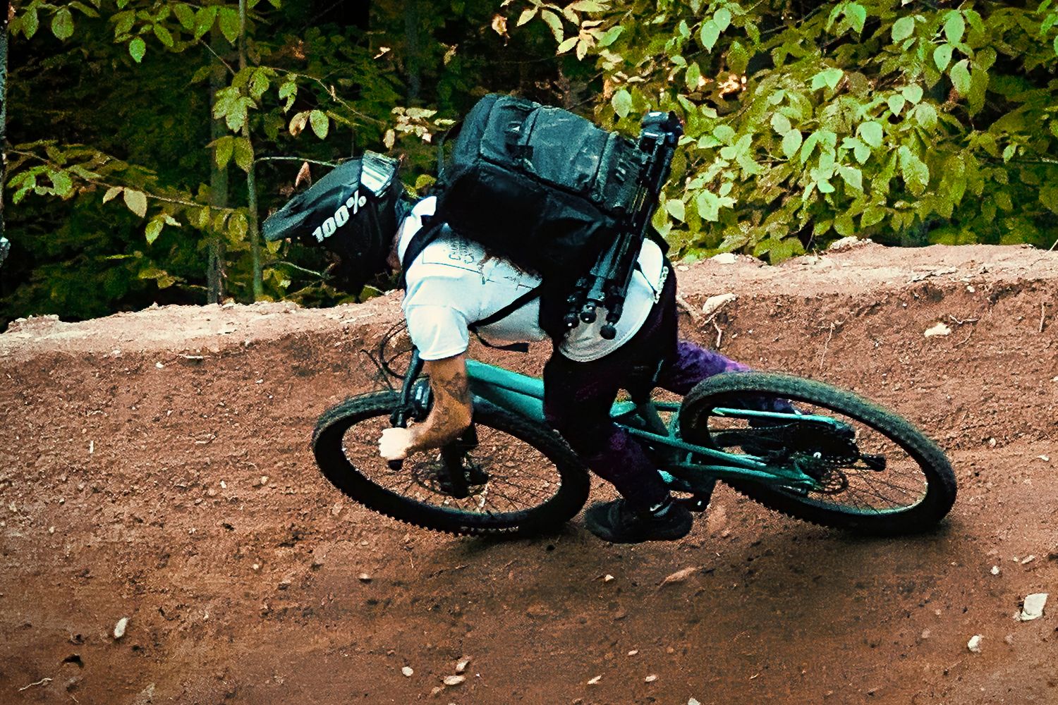 Mountain biker with large backpack navigating a dirt trail through a forest on a teal bike.