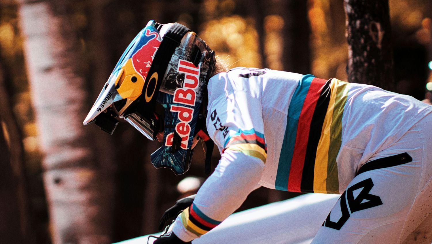 Red Bull athlete in rainbow world champion jersey and helmet rides mountain bike through forest trail.