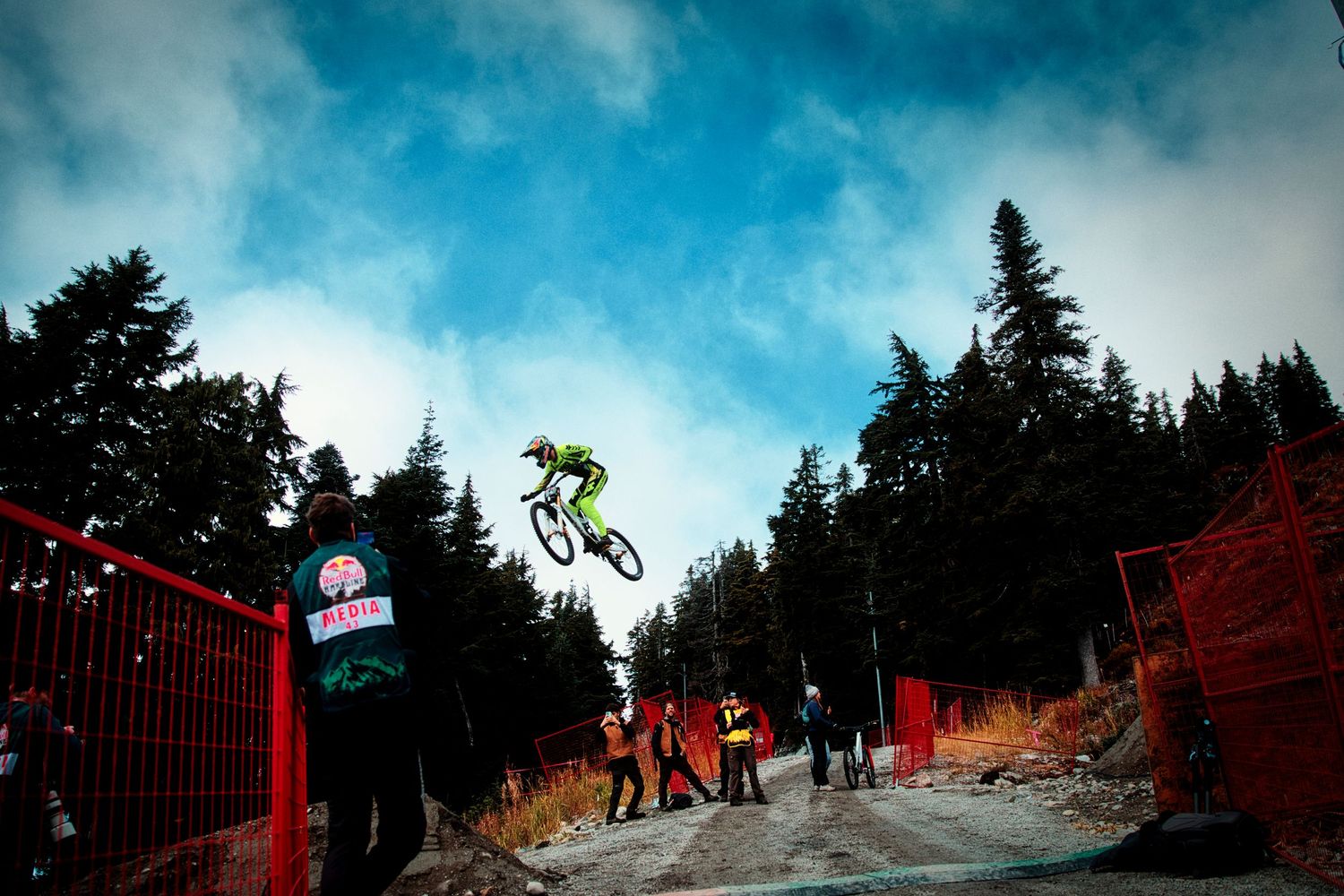 Downhill mountain biker in green gear soars off a dirt jump above spectators and a red fence, with evergreen forests behind.