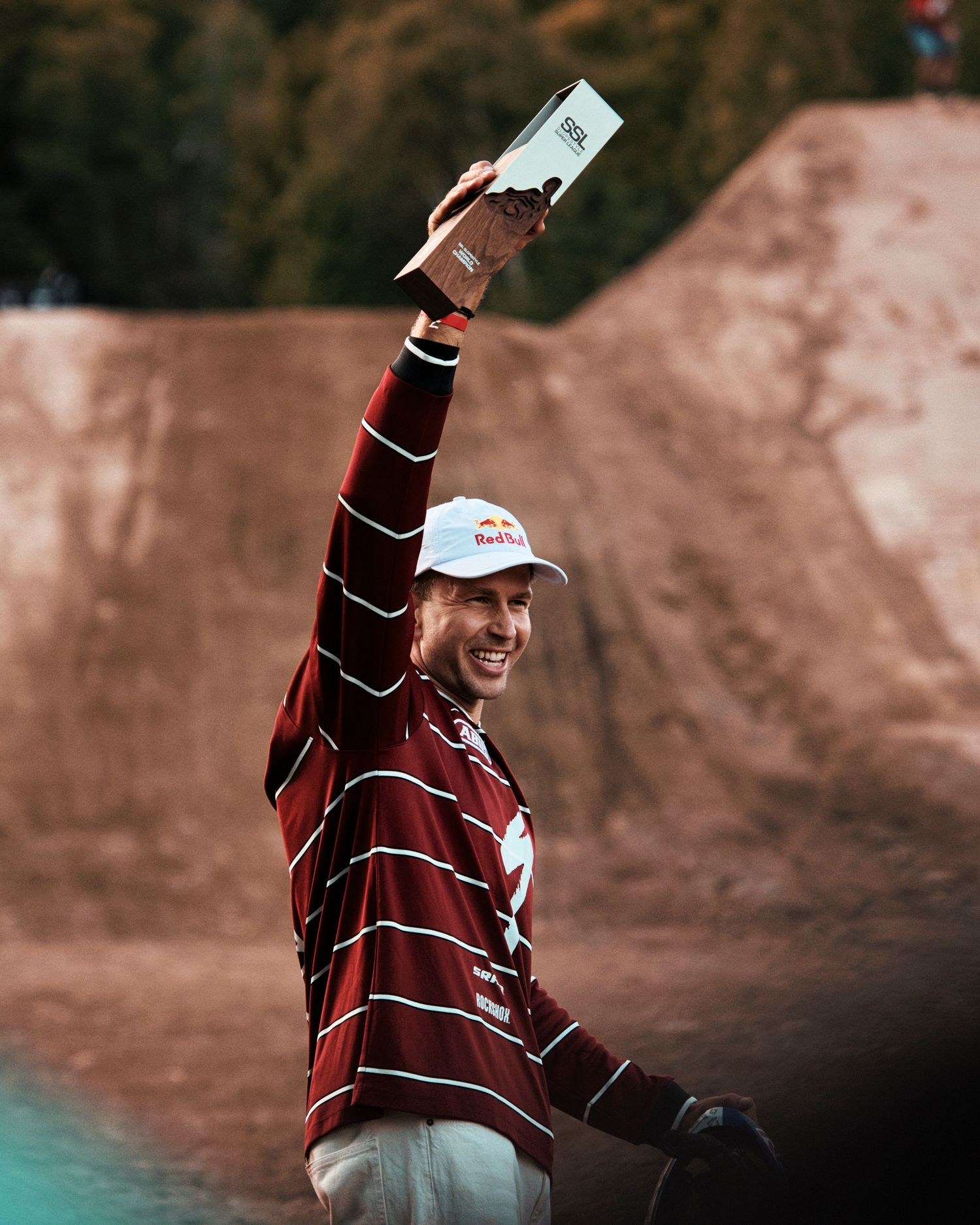 A smiling athlete in a Red Bull cap raises a trophy at a BMX dirt jump competition venue.