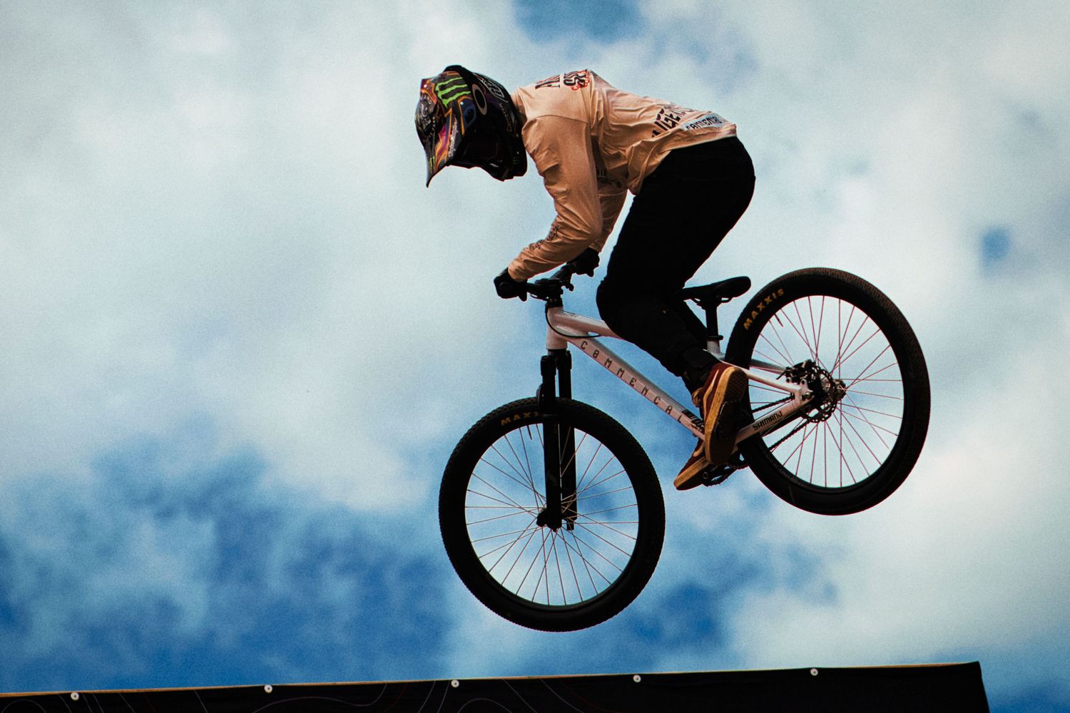 Slopestyle rider launching off a ramp performing a jump trick against a bright cloudy blue sky by Dave Clément, Québec.