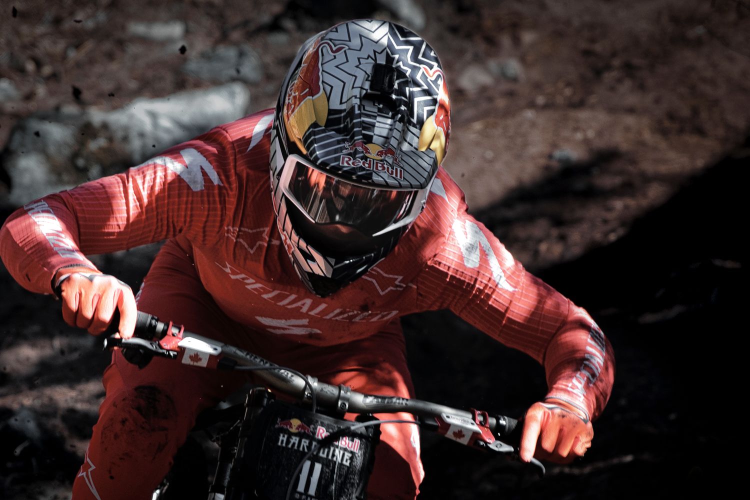 Mountain biker in a red racing suit and graphic full-face helmet gripping handlebars while riding a rocky downhill trail.