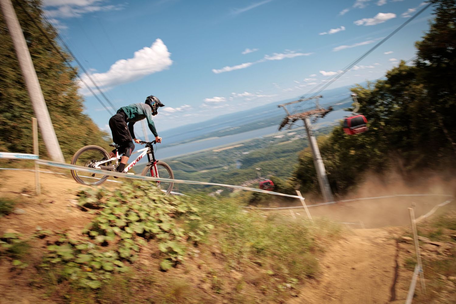 A downhill mountain biker descends a steep trail beside ski lifts with a scenic view below by Dave Clément, Québec.