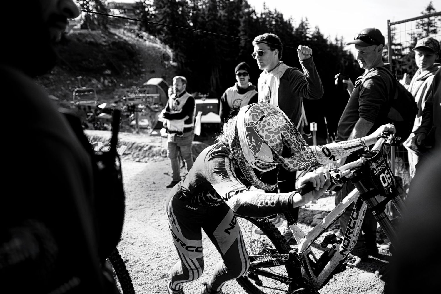 Black and white photo of a downhill mountain biker in a full-face helmet leaning over a bike as spectators watch on a dirt trail.