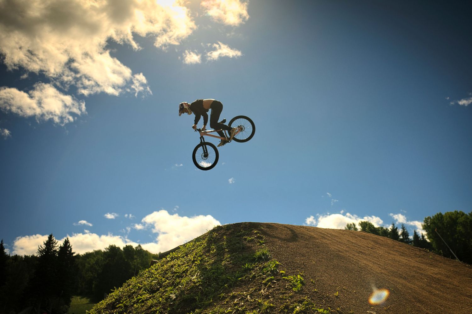 A mountain biker performs a high aerial trick off a dirt jump ramp against a blue sky with clouds.