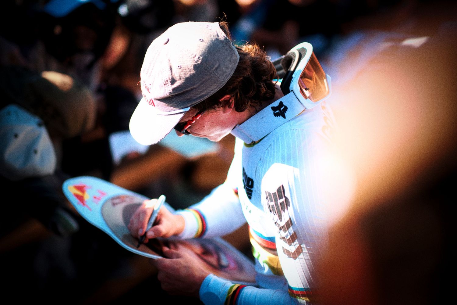 Cyclist in a gray cap, sunglasses and a white and blue racing kit signs an autograph on a poster in warm golden light.