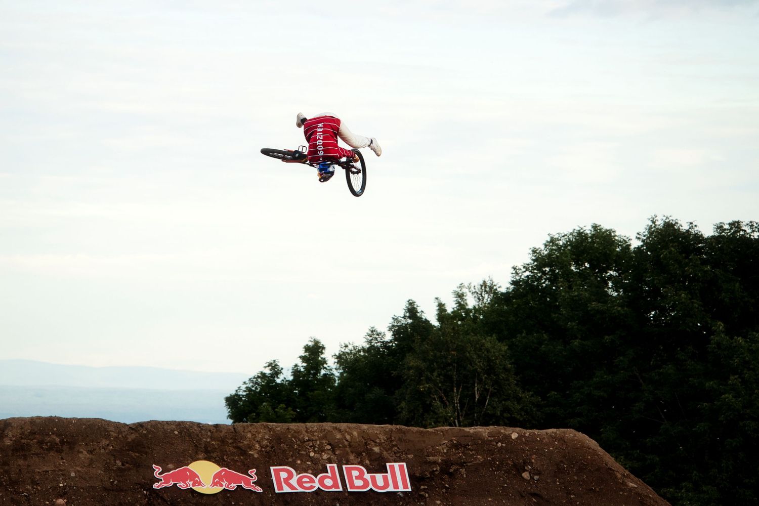 David Godziek soaring high above Red Bull branded dirt jump ramp against clear sky backdrop by Dave Clément, Québec.