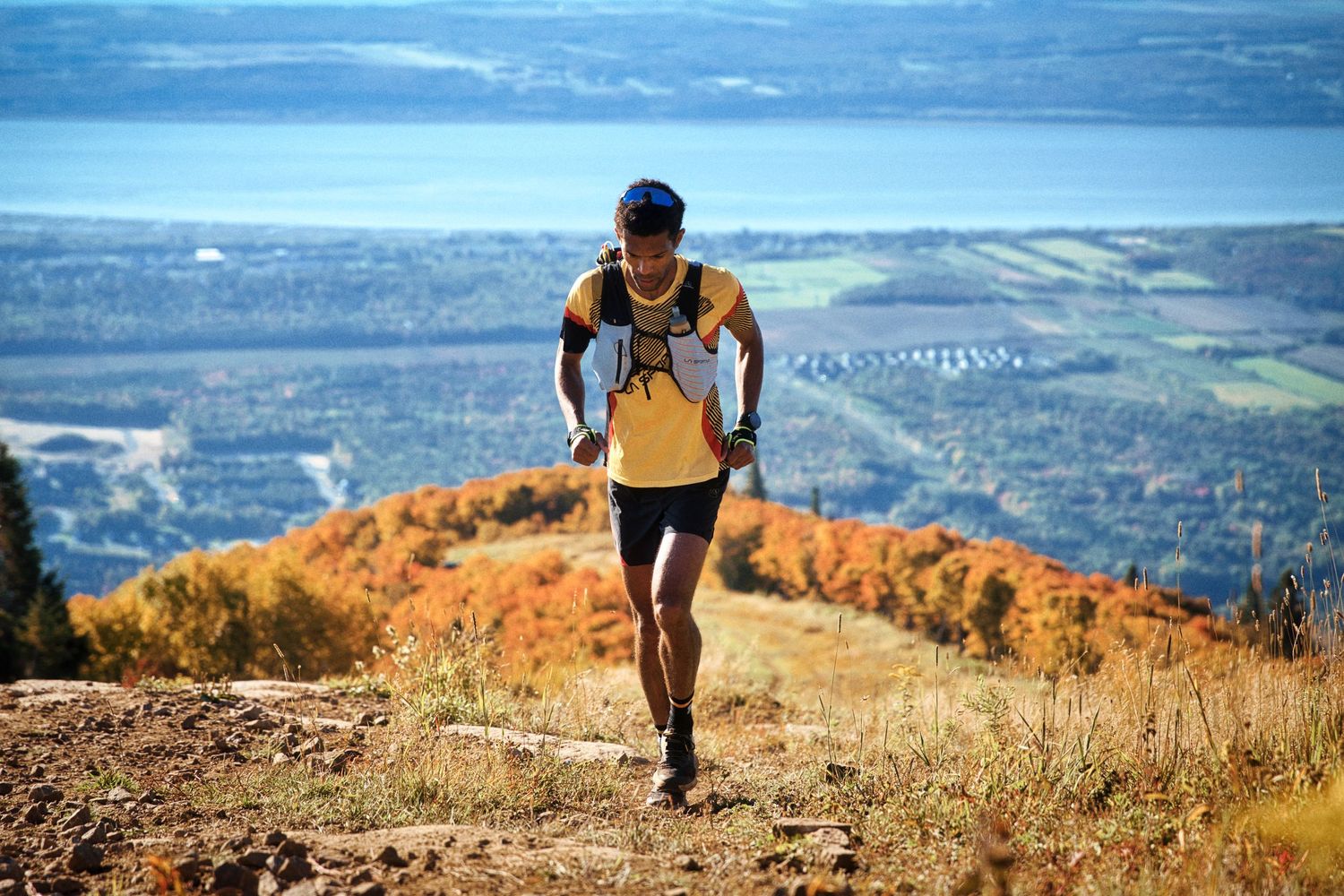 Trail runner in yellow shirt climbs rocky mountain path with colorful autumn valley and lake views behind.