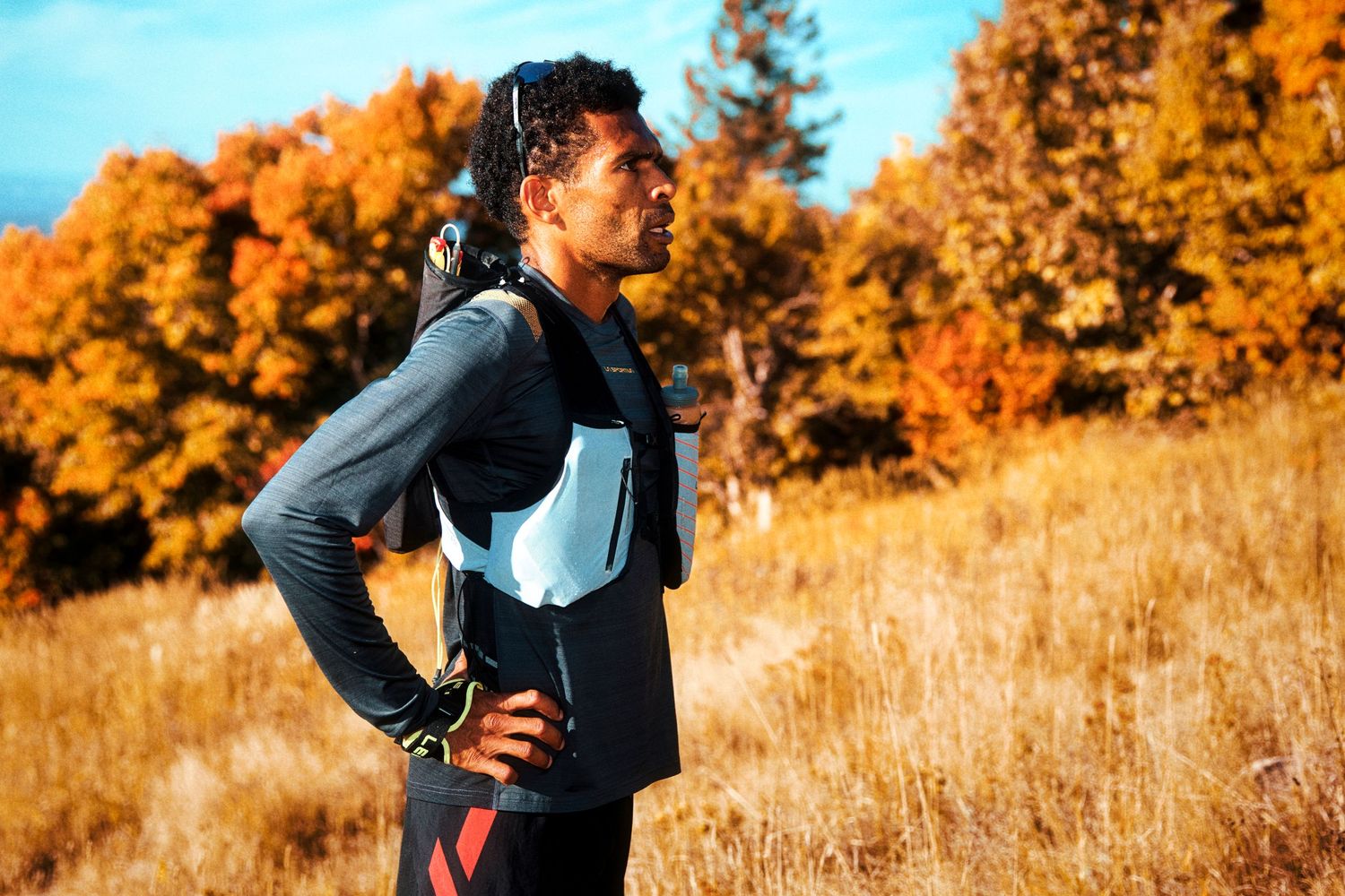 A male trail runner wearing a hydration vest pauses on a hillside trail surrounded by vibrant autumn foliage.