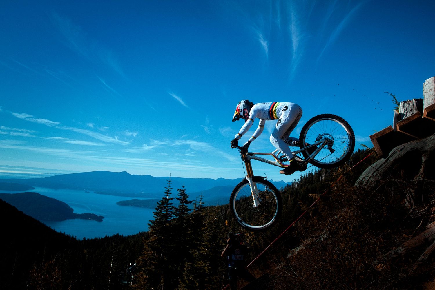 Mountain biker in a white jersey and helmet rides a steep rocky trail above a blue coastal inlet with forested mountains under a bright sky.