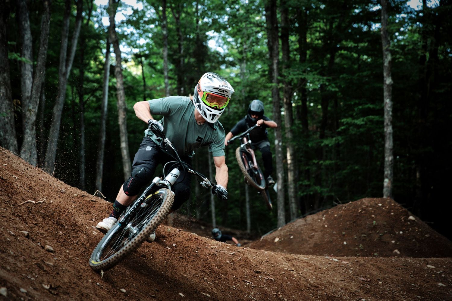 Two mountain bikers carve through dirt berms on a forested trail during air dh riding session by Dave Clément, Québec.