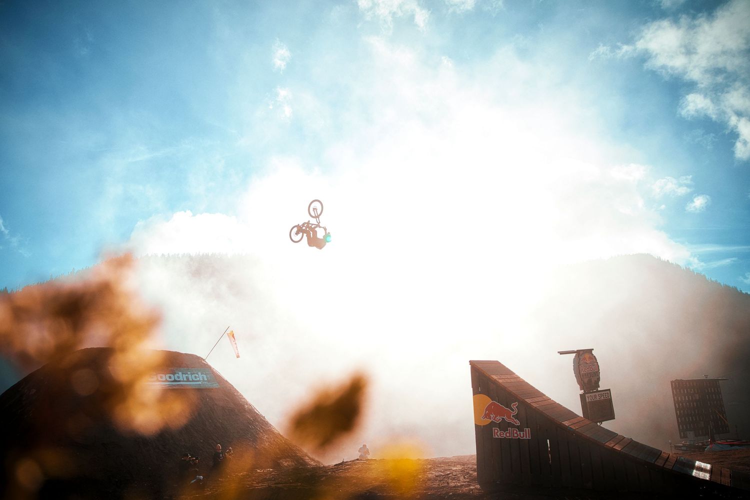 BMX rider soaring high above a dirt jump ramp against a bright sunlit sky during an extreme action sports event.