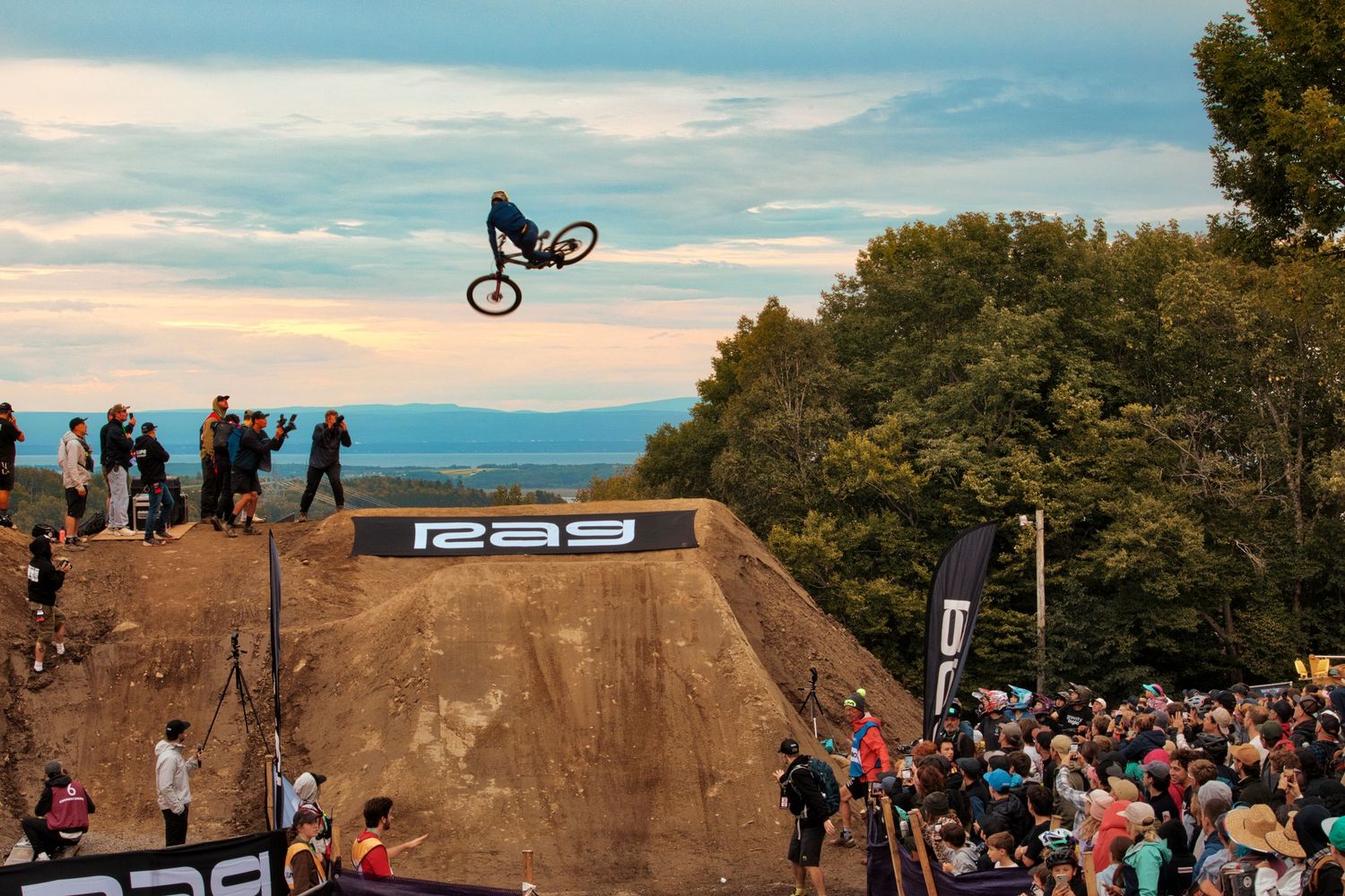 Mountain biker performing massive whip over dirt jump ramp before large outdoor crowd by Dave Clément, Québec.
