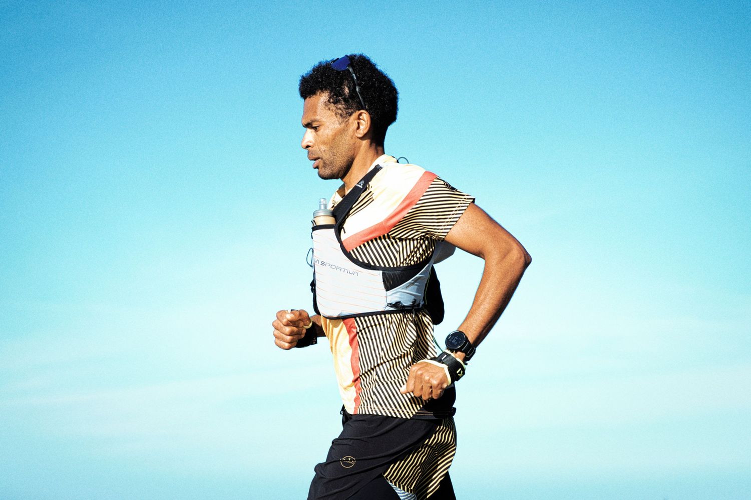 Male runner in athletic vest and shorts jogging against a clear blue sky outdoors.