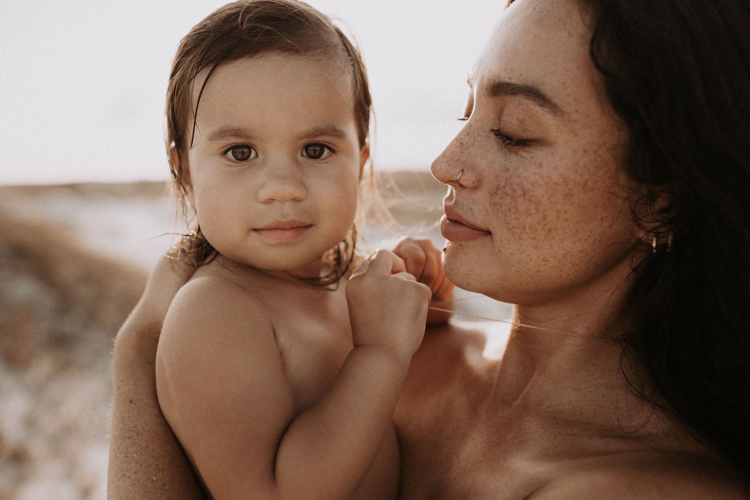 Freckled mother holding baby close, gazing lovingly at child in warm sunlight.