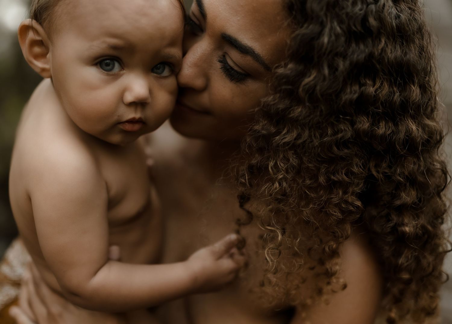 A mother with curly hair gently kisses her baby's cheek in a warm, intimate close-up portrait.