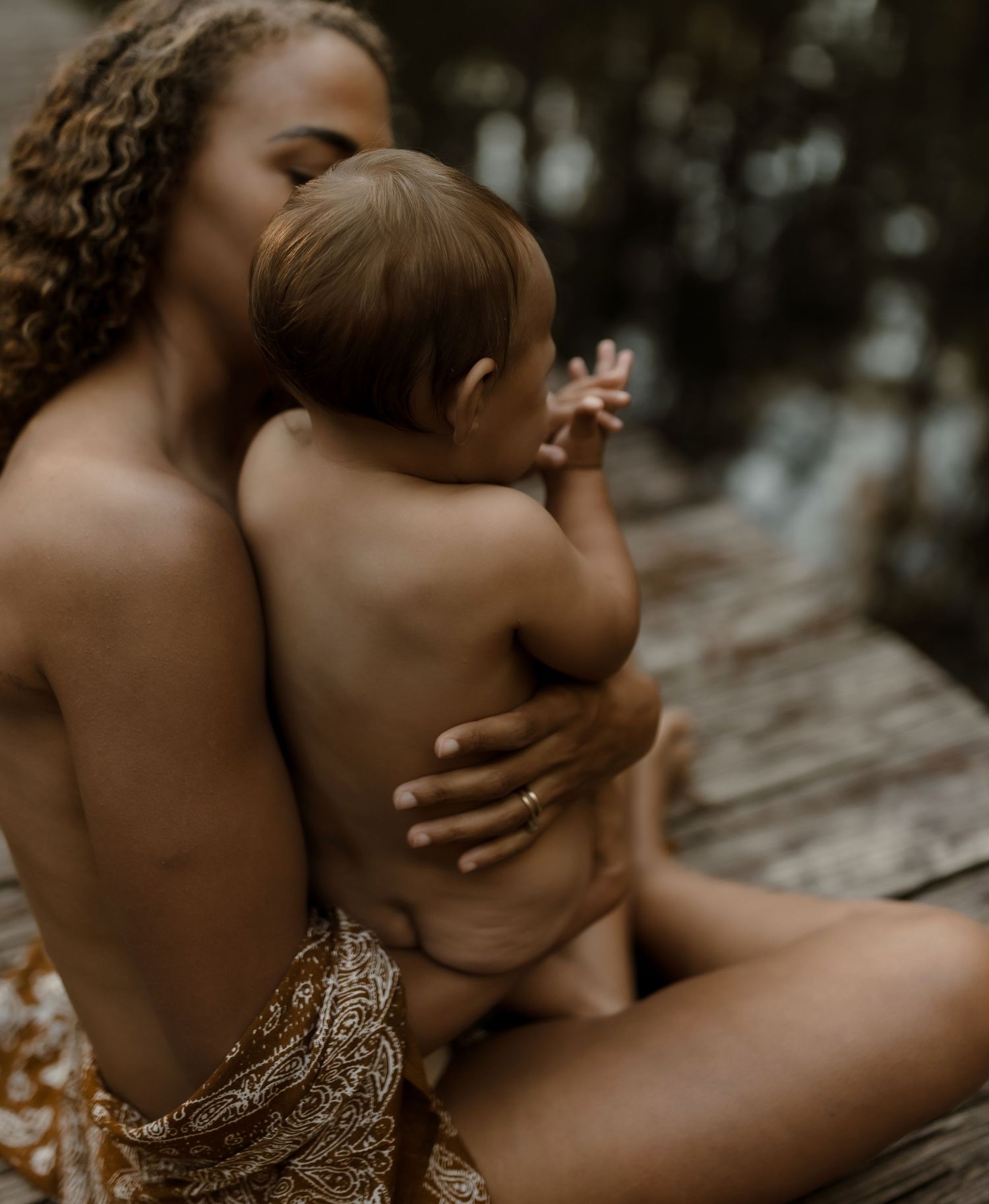 Mother tenderly embraces baby skin-to-skin while seated on rustic dock near calm water.