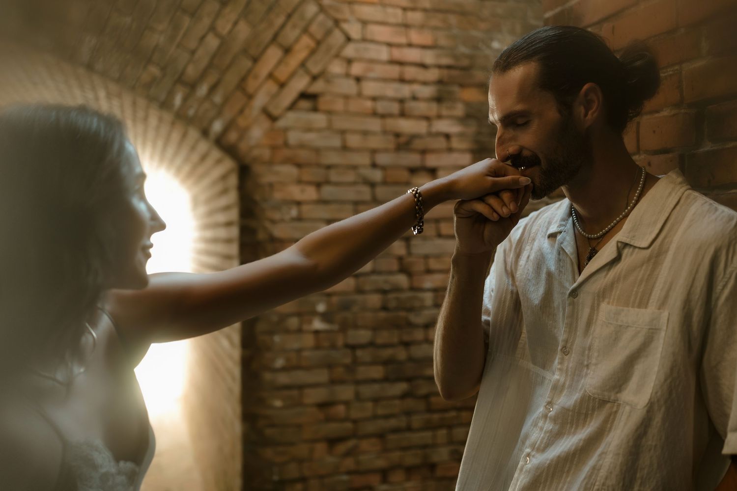 A man in a white shirt kisses a woman's hand near a brick wall in a romantic, dimly lit setting.