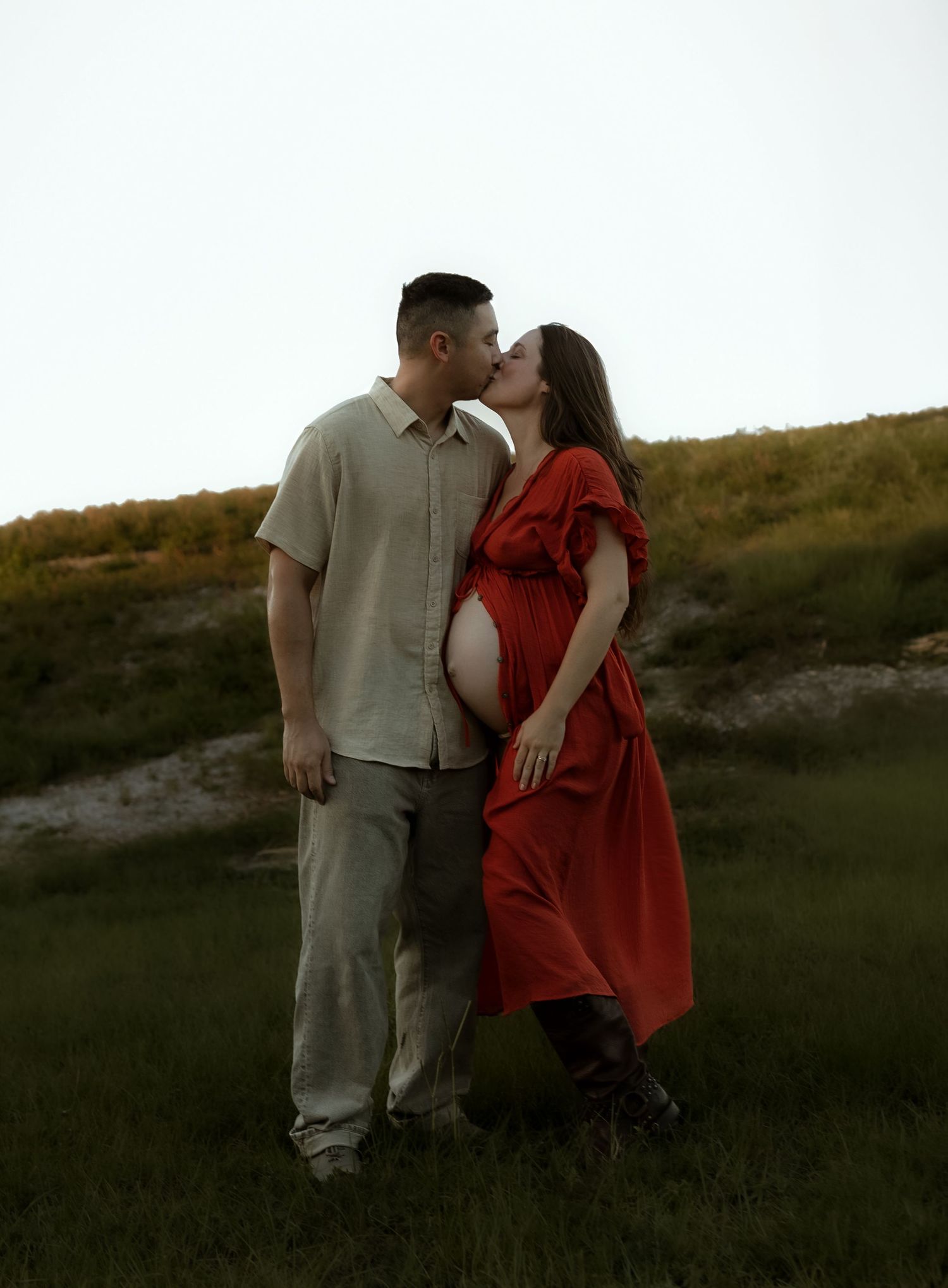 Pregnant woman in red dress kissing partner in beige outfit outdoors in green field at dusk.