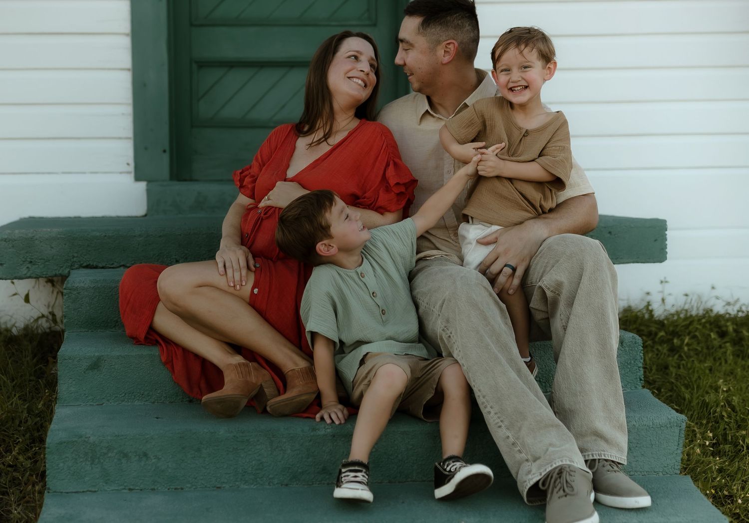 A happy family of four sits on green porch steps, smiling and laughing together in casual outdoor portrait.