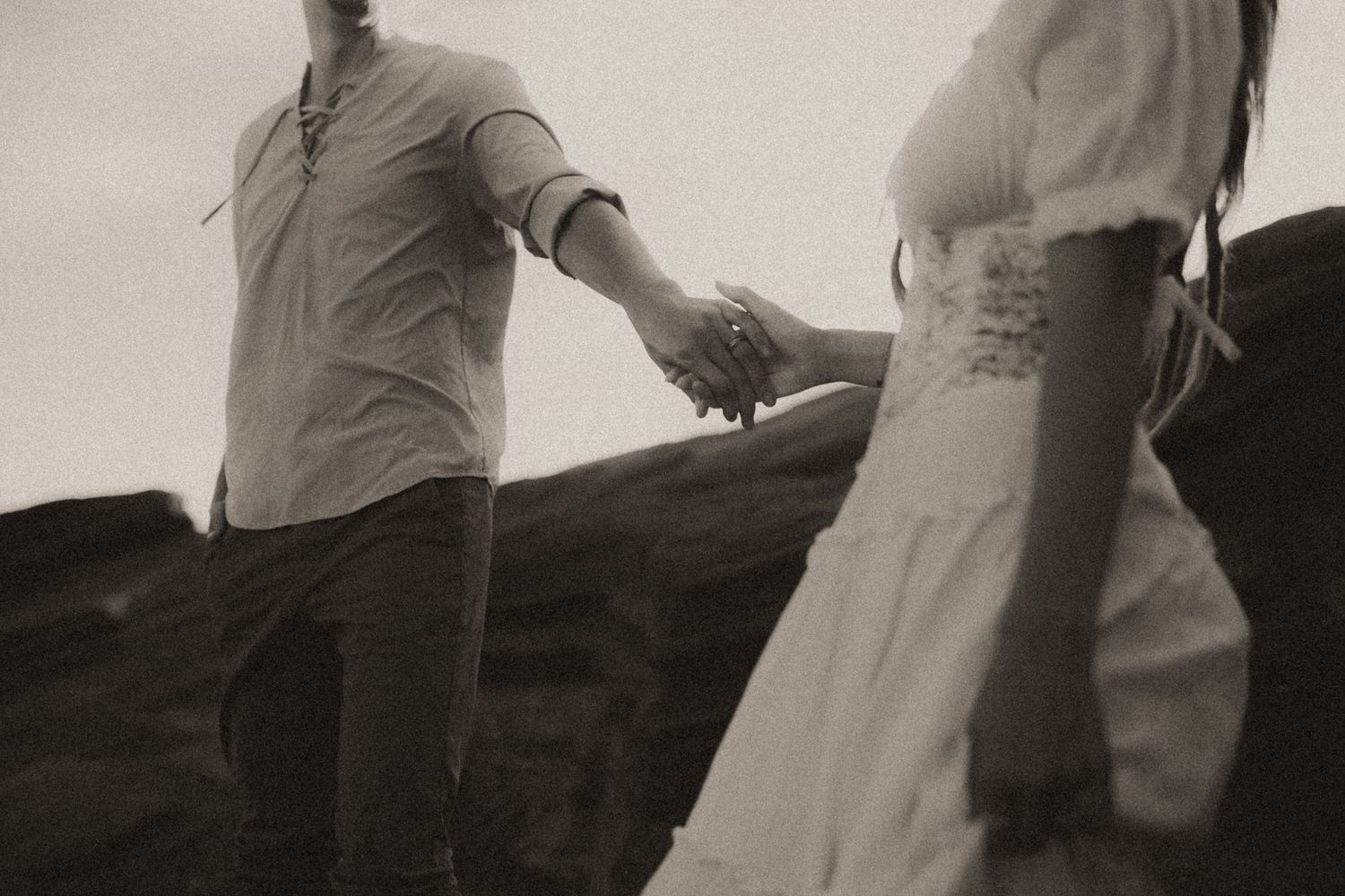 Black and white photo of a couple holding hands on rocky terrain, woman in white dress leading the way.
