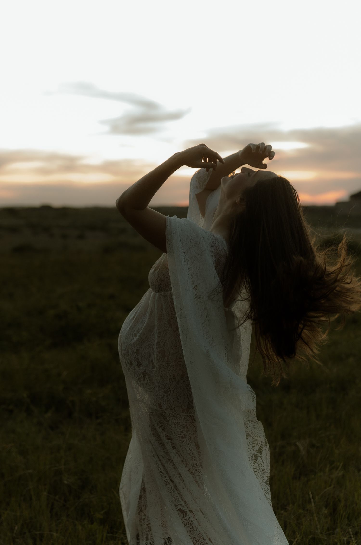 A silhouette of a pregnant woman in a white lace dress dancing in a field at sunset.
