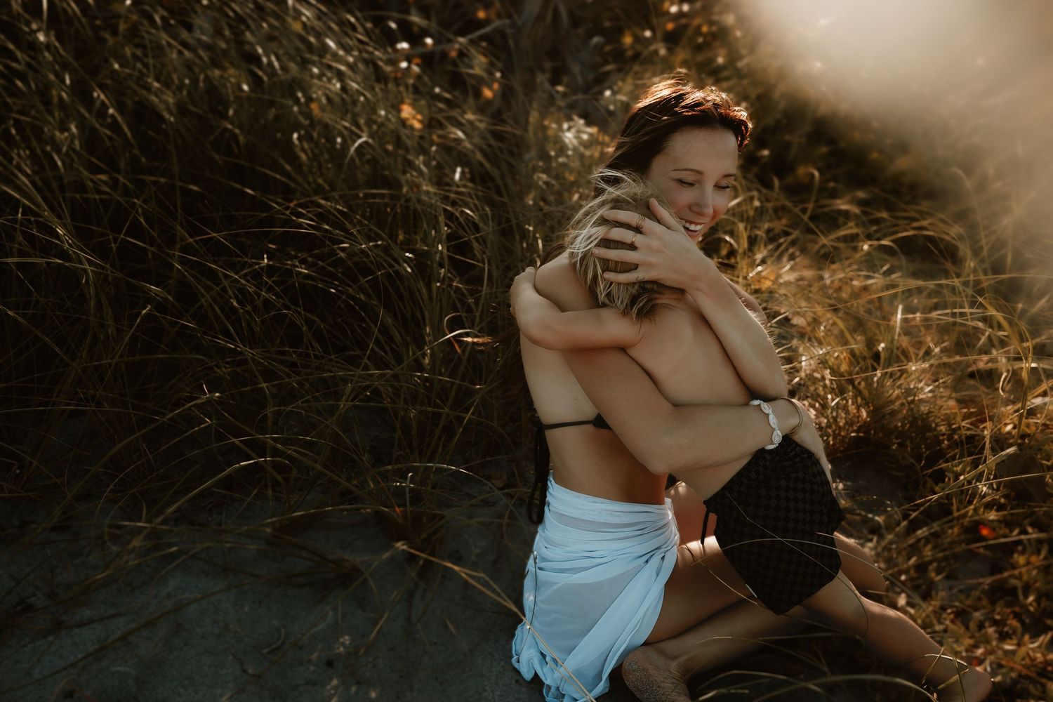 Woman smiling while hugging child in sandy dunes with golden grass.