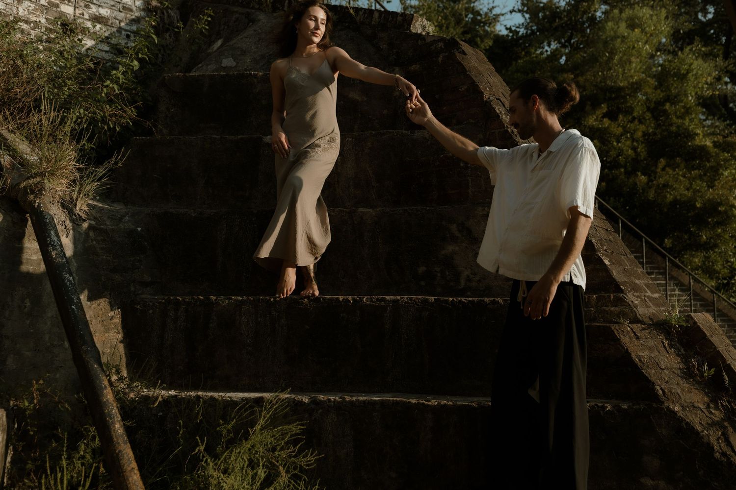 Couple holding hands on stone steps outdoors, woman in beige dress, man in white shirt, surrounded by lush greenery.
