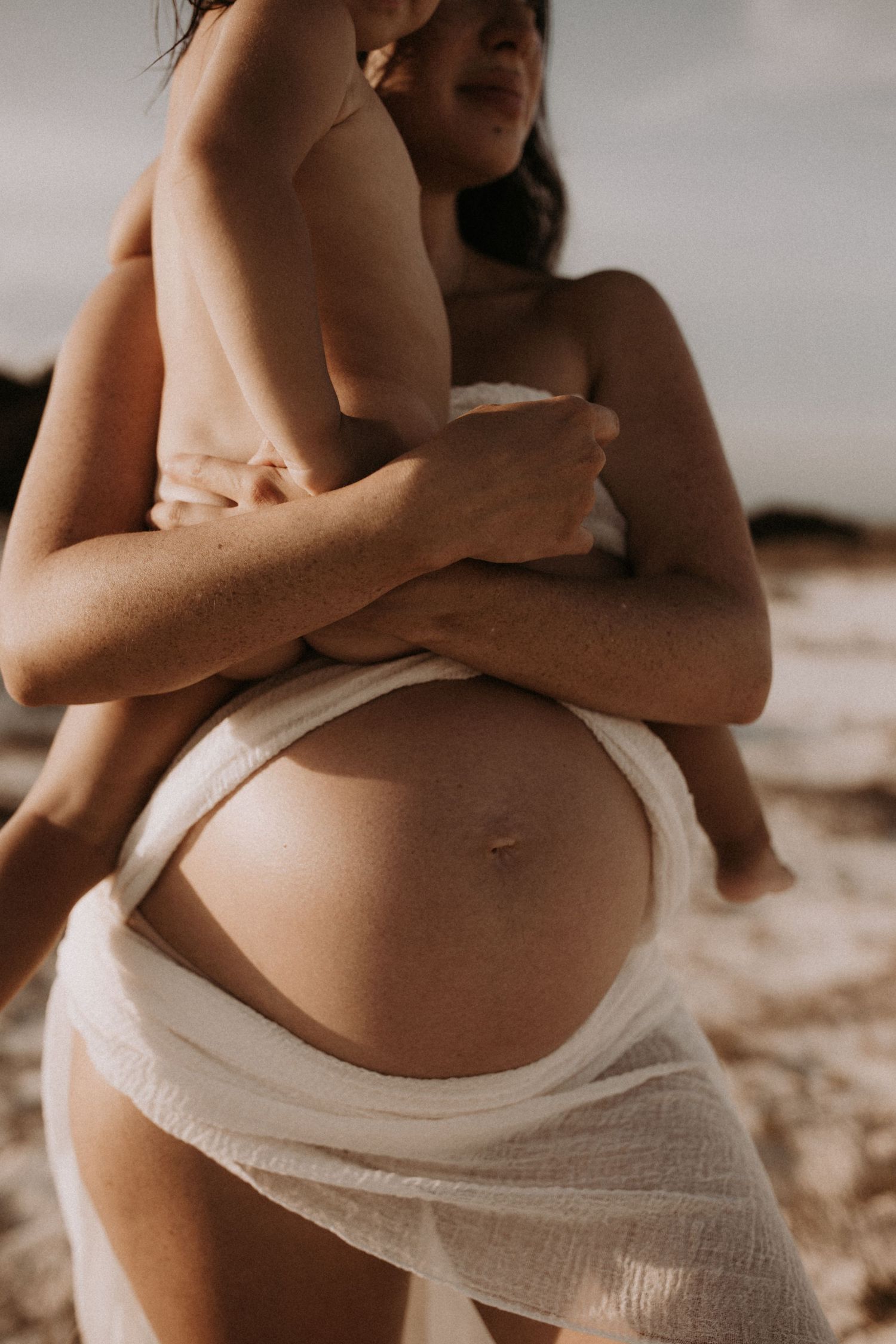 Pregnant woman holding toddler, bare baby bump visible, wrapped in white fabric outdoors.