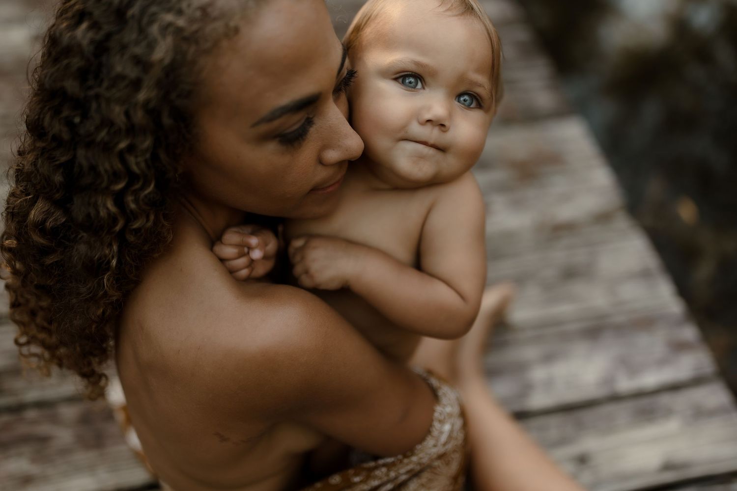 A mother with curly hair tenderly kisses her baby's cheek while holding the infant close outdoors.