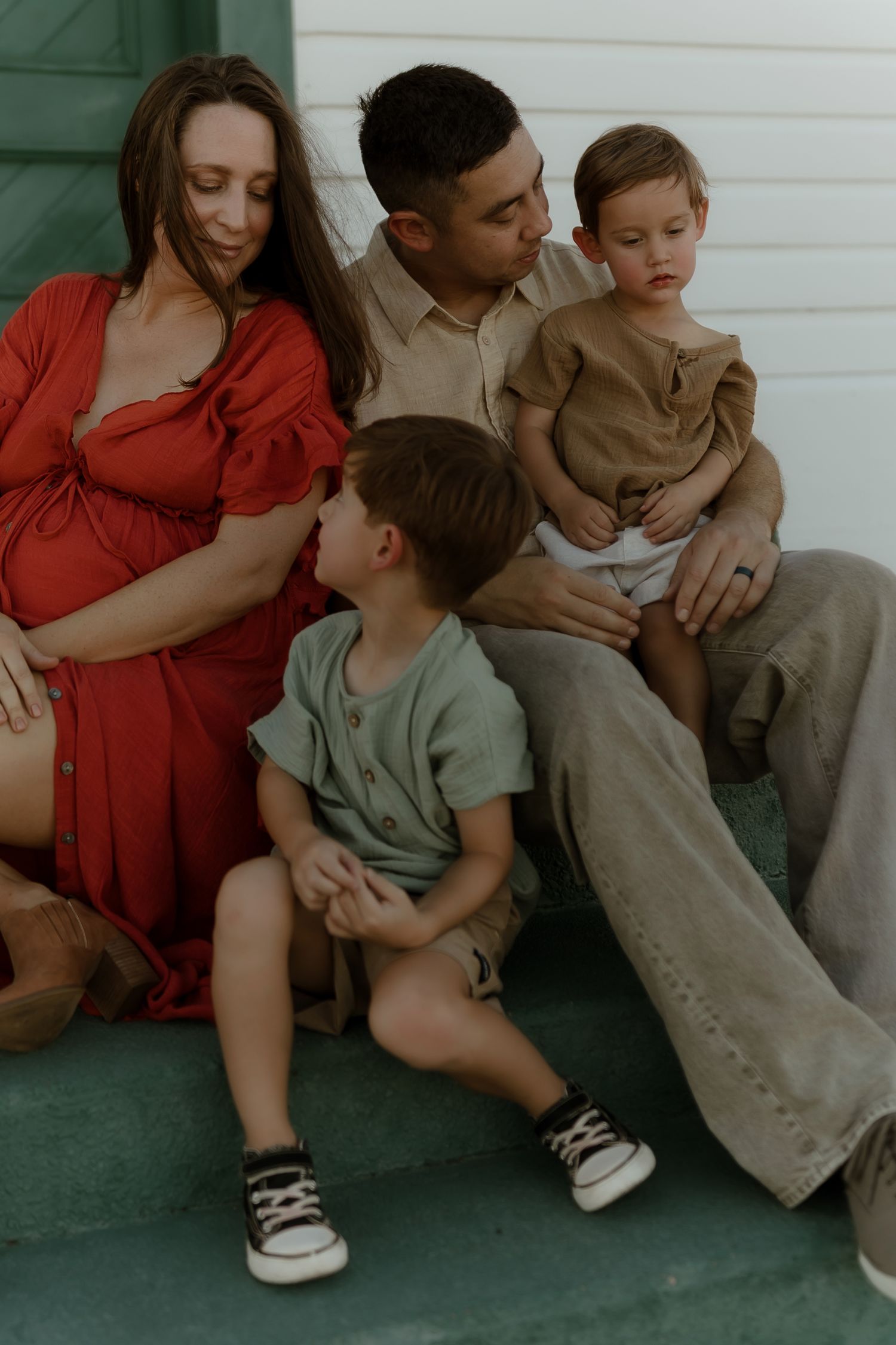 A pregnant woman in red dress sits on steps with her partner and two young toddler boys for a family portrait.